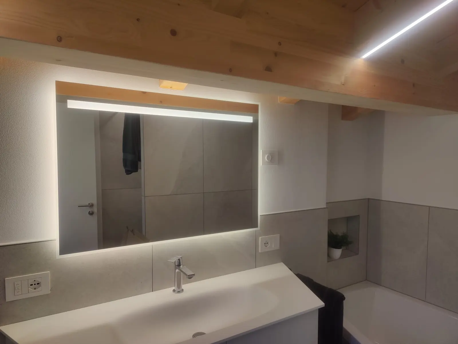 Rechteckiger Spiegel mit LED-Leiste im oberen Glasbereich und Hinterleuchtung, unter einer Holzbalkendecke.